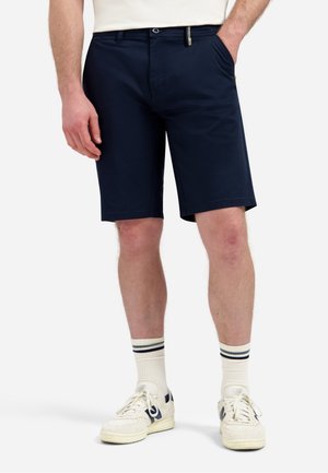 Man draagt donkerblauwe shorts, witte sneakers en witte sokken met zwarte strepen, staat met één hand in de zak.