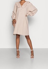 Vestido de color rosa pálido, con textura, cuello con volantes, mangas largas y detalles de botones, combinado con tacones nude con tira en el tobillo.