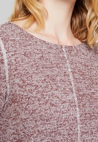 Suéter acogedor de punto texturizado en una mezcla de tonos rojos y blancos, con un escote amplio y costuras visibles. Tejido suave con un ajuste ligeramente holgado.