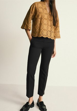 Vrouw staat met handen in de zakken, gekleed in een beige geperforeerde blouse, zwarte broek en zwarte loafers tegen een effen lichte achtergrond.