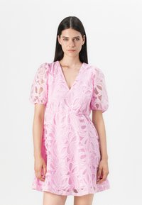 Abito in pizzo di un delicato rosa chiaro con maniche a sbuffo, scollatura a V e motivo floreale. Vita elastica e siluetta a trapezio con texture trasparente.