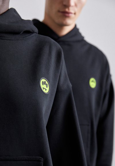 Black hoodies feature a front pocket and a circular green and black logo with a playful face on the left side. Soft fabric texture is visible.