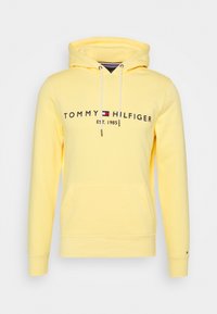 Sudadera con capucha amarilla de algodón con un bolsillo frontal, cordones y texto "TOMMY HILFIGER EST. 1985". Textura suave.