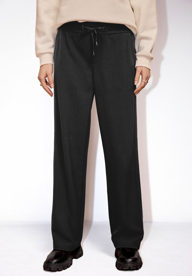 Personne portant un pantalon noir ample à taille ajustable avec cordon et des chaussures noires épaisses, debout devant un fond blanc.