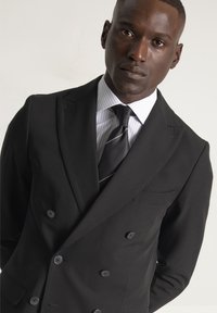 Chaqueta de traje negra de doble botonadura con solapas con muesca, con botones grises y un bolsillo en el pecho, combinada con una camisa de vestir a rayas y una corbata.