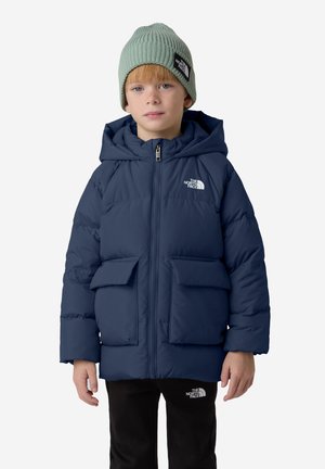 Chaqueta de plumas azul marino con capucha, que cuenta con dos bolsillos frontales y un logo. El niño lleva un gorro de punto verde claro. Pantalones negros.