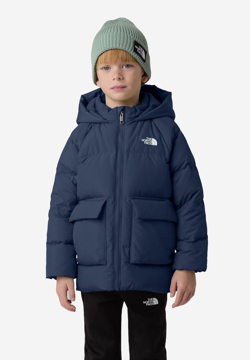 Námorně modrá péřová bunda s kapucí, obsahující dvě přední kapsy a logo. Dítě má na sobě světle zelenou pletenou čepici. Černé kalhoty.