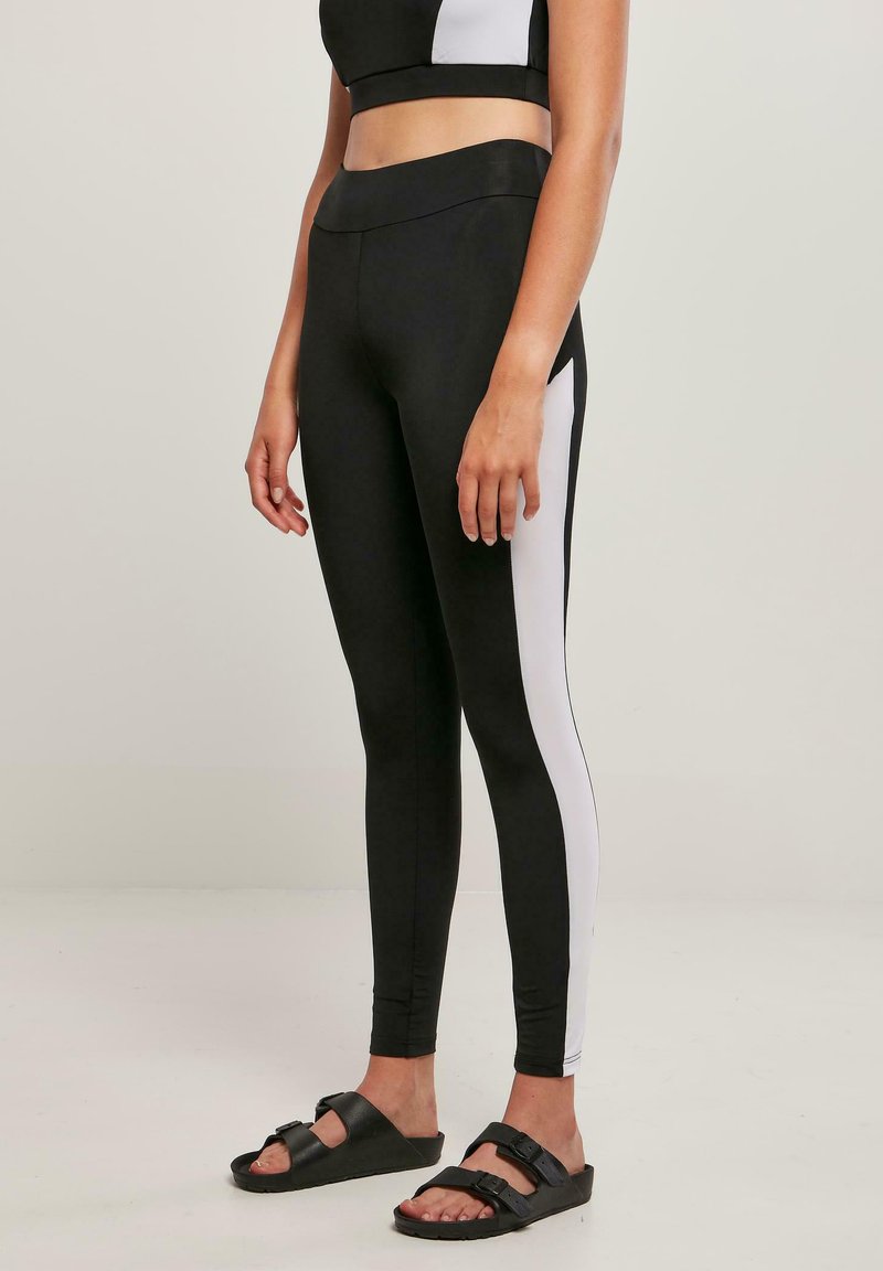 Leggings noirs à taille haute, avec des panneaux latéraux blancs. Confectionnés en tissu extensible, conçus pour le confort et la mobilité, assortis à des sandales noires.