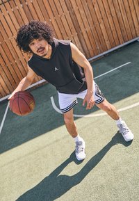 Svart ermeløs hettegenser med hvit logo, hvite shorts med svarte striper, og grå basketballsko. Holder en basketball på en bane.