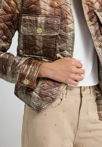 Quiltet jakke i brun og creme med et geometrisk mønster, der har to forlommer og guldknapper som accent. Sat sammen med beige bukser udsmykket med gulddutter.