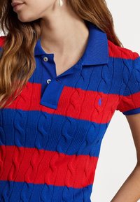Pruhovaná polo tričko v červené a modré barvě, s texturovaným vzorem pletené kabely. Má modrý límec a tříknoflíkovou légu. Malé logo na hrudi.