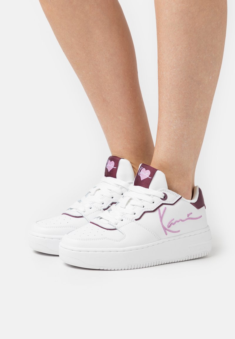 White sneakers with a sleek design, featuring a maroon accent at the collar, purple signature detail on the side, and textured rubber sole.
