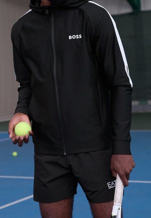 Person in schwarzer Sportbekleidung hält in der einen Hand einen Tennisball und in der anderen einen Tennisschläger auf einem blauen Tennisplatz.