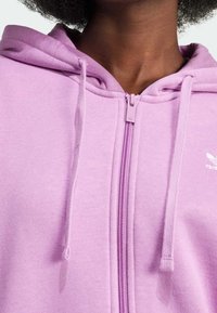 Osoba v světle fialové mikině na zip s kapucí na stažení a malým bílým logem Adidas na hrudi.