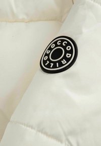 Czarnobiały, okrągły nadruk logo "Coccodrillo" na białej, puchowej powierzchni materiału, prawdopodobnie część kurtki lub płaszcza.