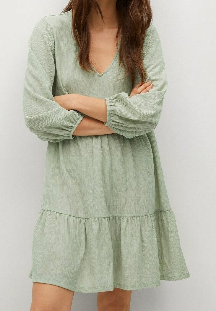 Femme portant une robe vert clair texturée à manches longues avec un col en V et un ourlet à volants, les bras croisés sur la poitrine.