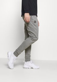 Graue Sport-Jogginghosen mit elastischen Bündchen, Seitentaschen und einem Under Armour Logo-Patch, kombiniert mit weißen Sport-Schuhen und Socken in Knöchellänge.