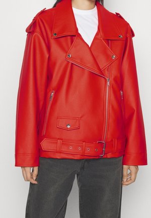Rote Bikerjacke mit silbernen Reißverschlüssen, Druckknöpfen und Gürtel, getragen über einem weißen Hemd und dunklen Hosen an einer stehenden Person.