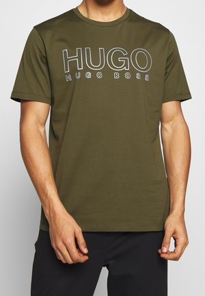 Mężczyzna noszący oliwkowozielony T-shirt z krótkim rękawem z dużym białym napisem „HUGO” i mniejszym „HUGO BOSS” na klatce piersiowej.
