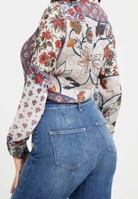 Kvinna som bär högmidjade blå jeans och en långärmad blommönstrad blus, sedd bakifrån mot en vit bakgrund.