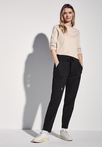 Top de manga larga beige combinado con pantalones negros de cintura elástica que cuentan con dos bolsillos frontales. El modelo lleva zapatillas blancas y está de pie frente a un fondo liso.