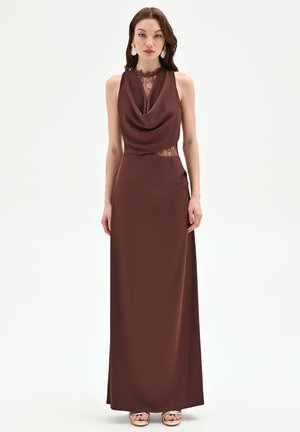 Donna in piedi che indossa un abito lungo senza maniche marrone scuro con dettagli in pizzo e orecchini dorati su uno sfondo chiaro semplice.