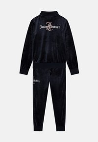 Črni velur trenirka, ki vključuje jakno z zadrgo, na hrbtni strani z napisom "Juicy Couture" v kamenčkih in usklajene hlače z oprijetimi nogavicami ter podobnim logotipom.