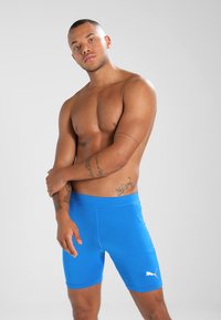 Blå atletiske shorts lavet af strækkeligt stof. Har en glat tekstur, tætsiddende design og hvid Puma-logo på den nederste side.