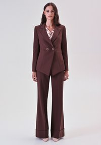 Braunes maßgeschneidertes Anzug mit einem doppelläufigen Blazer, der runde metalische Akzente aufweist, und einer weiten Hose, kombiniert mit einer blusen in Cream mit Punkten.