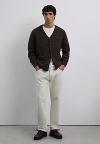 Hombre de pie con las manos en los bolsillos, vestido con un cárdigan oscuro de botones marrón, camisa blanca, pantalones beige claros y zapatos marrón oscuros, sobre un suelo gris.