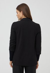 Donna con capelli castani lisci che indossa un blazer nero con le maniche arrotolate, di spalle contro uno sfondo chiaro e semplice.