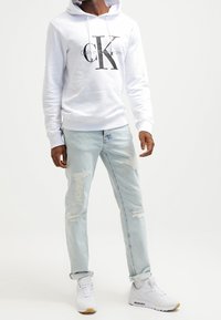 Man wearing white Calvin Klein hoodie, light distressed jeans, and white sneakers, standing against plain white background.