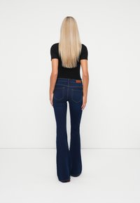 Mujer rubia vestida con una blusa negra de manga corta y unos vaqueros de campana azul oscuro, de pie con la espalda hacia el espectador contra un fondo blanco.