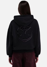 Schwarzer Kapuzenpullover mit einem auffälligen lila Logodesign auf dem Rücken, aus weichem Stoff mit entspannter Passform und gerippten Bündchen.