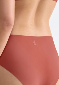 Primo piano della parte bassa della schiena e della vita di una persona che indossa biancheria intima senza cuciture di un rosso-marrone lucido, con un piccolo logo "Sloggi" sulla parte posteriore.