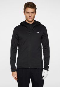 Svart zip-up hoodie med en tight passform och huva, tillverkad av textilmaterial. Har en liten vit logga på bröstet och framfickor med dragkedja.