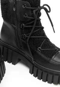 Botín negro hasta el tobillo con parte superior de cuero texturizado y tela esponjosa, con suela gruesa de goma, diseño con cordones y cremallera lateral.