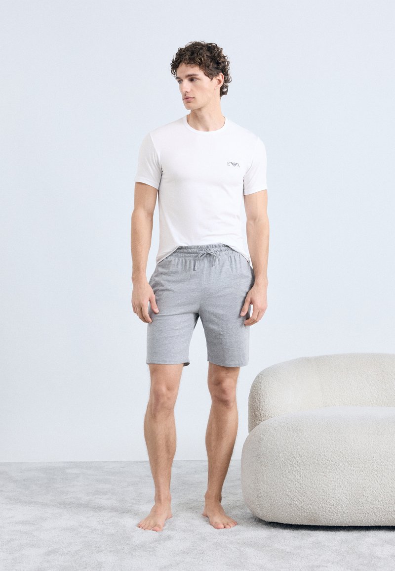 Balta īsrokas krekls ar logotipu, kopā ar pelēkām šortām ar elastīgu jostu. Vienkāršs dizains, gluda tekstūra, stāv uz gaiša paklāja.