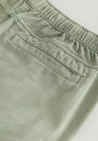 Pantalon à taille élastique vert clair avec tissu texturé et une poche passepoilée arrière, présenté en gros plan.