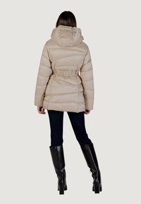 Giacca a vento beige con cappuccio, vita cinchata e texture a coste. Indossata sopra jeans scuri e stivali neri al ginocchio, vista da dietro.