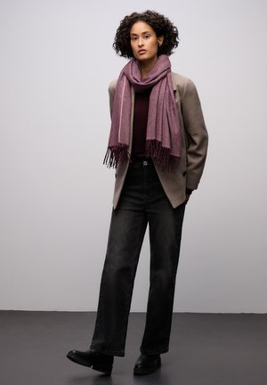 Écharpe tissée violette à franges, portée sur un haut bordeaux et un blazer gris, associée à un jean noir taille haute et des bottines noires.