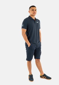 Camisa polo marina de manga corta con ribete blanco; combinada con pantalones cortos de carga marinos. Usada con zapatillas marinas que tienen acentos naranjas.