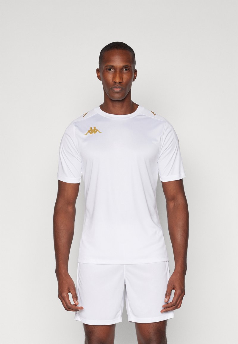 Camiseta atlética blanca hecha de una tela suave, con mangas cortas, cuello redondo y acentos dorados en los hombros y el logo. Pantalones cortos blancos a juego.