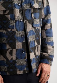 Wool flannel shirt jacket with a blue, gray, and black geometric pattern. Features button front, two chest pockets, and a textured finish.