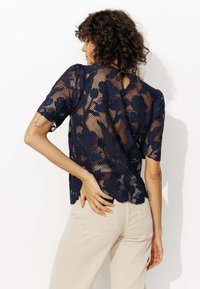 Haut en dentelle bleu marine avec des motifs floraux et des manches courtes, présentant un design en maille et une légère ouverture dans le dos, associé à un pantalon de couleur claire.