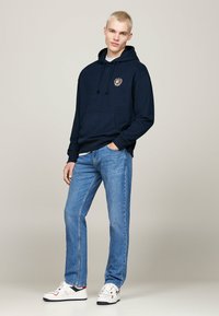Felpa con cappuccio di colore blu navy con tasca frontale e emblema, abbinata a jeans in denim azzurro chiaro e sneakers bianche con dettagli rossi e blu navy.