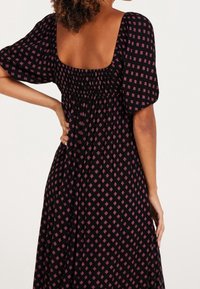 Kvinna som bär en svart klänning med rosa diamantmönster, puffärmar och en låg fyrkantig rygg, stående med en hand på höften.