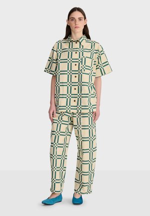 Jeune femme portant une chemise à manches courtes et un pantalon à carreaux crème et vert assortis, avec des chaussures bleues, debout devant un fond uni.
