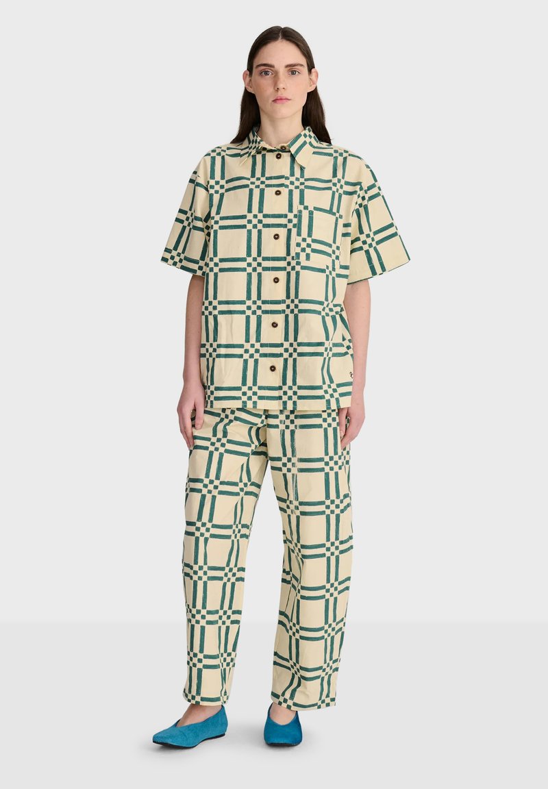 Jeune femme portant une chemise à manches courtes et un pantalon à carreaux crème et vert assortis, avec des chaussures bleues, debout devant un fond uni.