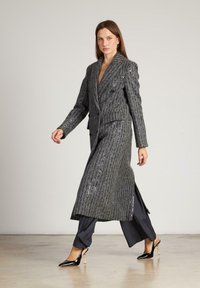 Abrigo largo de rayas grises con un sutil brillo, diseño ajustado, solapa de muesca, botones dobles y aberturas laterales. Lleva puesto sobre pantalones oscuros.